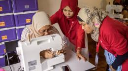 three women sit around a sewing machine wearing headscarves.