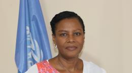 A woman in a white and pink shirt stands in front of a blue UN flag.
