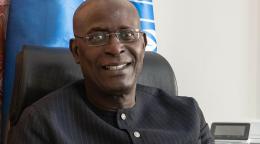 A man in a black suit and glasses sits in a chair with a blue UN flag behind him.