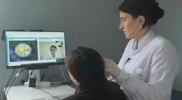 A doctor in a white coat diagnoses a woman in black clothing while another doctor is on a computer screen in front of them.