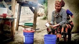 A woman sits wistfully and washes a metal pan/dish over buckets of water and soap.