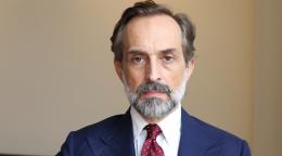 A man in a dark blue suit and red tie and pocket square sitting against a beige background