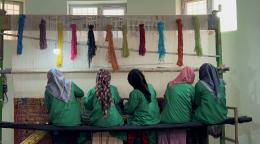 Women engaging in weaving, their heads are covered and their backs are turned to the camera