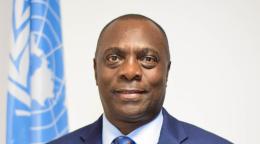 A man in a blue suit and blue tie stands in front of a blue UN flag