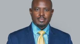 A man in a blue shirt, blue blazer and yellow tie against a grey background.