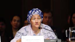 A woman in a lavender dress and deep purple headscarf speaks into a microphone