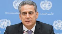 Muhannad Hadi sits in front of a lectern wearing a black suite and grey tie and behind him is a screen with UN logos