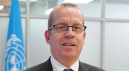 Un homme portant des lunettes et un costume sombre se tient devant un drapeau bleu portant le logo de l'ONU.