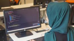 Фотография со спины женщины с покрытой головой, работающей с кодом в текстовом редакторе.