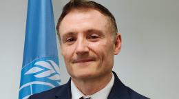 A man in a blue suit stands in front of a blue UN flag