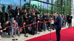A man in a black suit stands at a podium on a red carpet and addresses a group of mediapersons with cameras and microphones