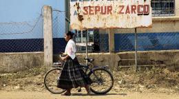 An indigenous woman walks her bicycle, there is a sign in the background and it is a dirt road. 