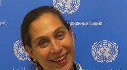 A woman in a white dress and colourful necklace leans against a blue background