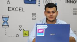 A boy is seated behind a laptop and looking at the camera. He has a colourful background behind him