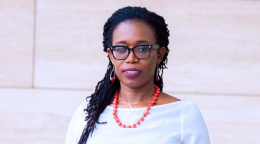 A woman is wearing a white top and orage necklace and glasses. She is looking at the camera and smiling