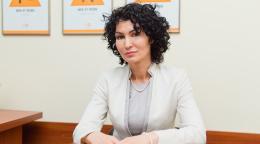 A woman in a white suit and black curly hair sits at a brown wooden desk with her arms crossed