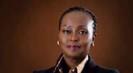 A woman in a black suit and golden brown scarf with her arms crossed against a brown background