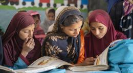 Three girls in red and brown dresses read over school textbooks in a classroom.