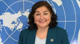 A woman with dark brown hair wearing a bluish-green suit and a white shirt standing in front of a blue wall with the emblem of the UN- white coloured states on a blue background,