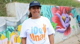 A woman in a colourful white shirt abd a cap stands in front of a painted wall.