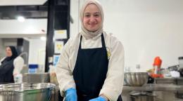 A woman in a white headscarf, black apron and blue gloves cuts vegetables on a kitchen counter.