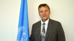 A man in a dark suit and a multicoloured tie stands next to a blue flag with the UN logo on it