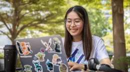 A girl in a white shirt wearing glasses smiles as she types into a laptop computer covered with stickers