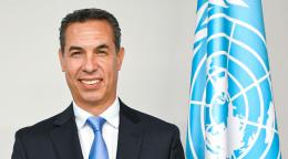 A man in a black suit and blue tie stands and smiles in front of white wall with a blue flag, the UN flag.