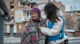 一位女性工作人员（右）搀扶着一位戴着紫色帽子裹着格子围巾的老年女性，站在被战乱破坏的房子前