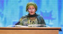 A woman in a green dress and headscarf speaks at a podium