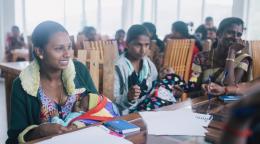 A row of women with children sit at desks while they fill out papers