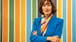 A woman in a blue suit and orange coloured scarf stands with her arms folded against a colourful wall.