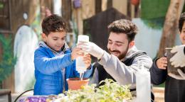 A man and a child hold a bottle of water to water a little plant in a container.