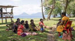 A group of people are gathered sitting on the ground, in front of a woman sitting on a chair talking to them
