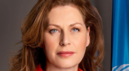 A woman with brown hair and a red shirt stands next to a blue UN flag