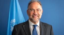 A man in a dark suit and blue tie, wearing spectacles (the RC for the Kosovo team) stands in front of a blue wall and a blue UN flag.