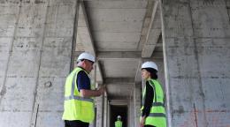 Two people are standing in front of a large infrastructure project speaking to each other 