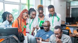 A group of men and women gather around a computer and appear in deep discussion.