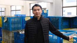 Retrato de Ying Changbin en su planta de producción de utensilios de cocina, rodeado de materiales y componentes.