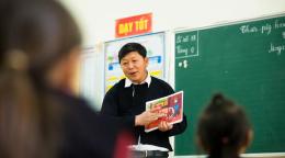 An image of a teacher pointing out something in a textbook in front of a class of students.