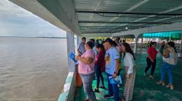 Un grupo de personas, de pie en una pasarela cubierta, observa el mar mientras sostiene cuadernos en las manos.