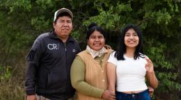 Tres personas sonríen juntas al aire libre frente a unos árboles.