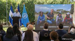 Un hombre da un discurso ante un público, junto a banderas y una pantalla con una imagen sobre trabajo agrícola.
