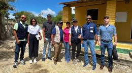 Representantes de organizaciones internacionales y autoridades locales visitan una comunidad rural frente a un edificio comunitario.