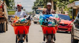Two street vendors push red wheelbarrows filled with assorted footwear and goods along a busy road lined with vehicles and buildings.