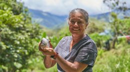 Una mujer sonriente sostiene un fruto entre las manos en un entorno rural montañoso y rodeado de vegetación.