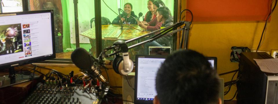 Four young people speak into a microphone in a green room in front of a person with 