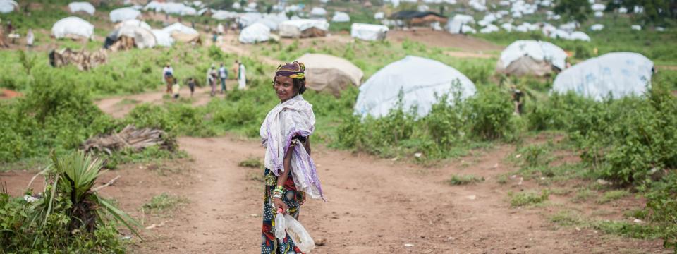 A woman turns back and smiles at the camera as she walks towards a green patch, with tents dotting the horizon.