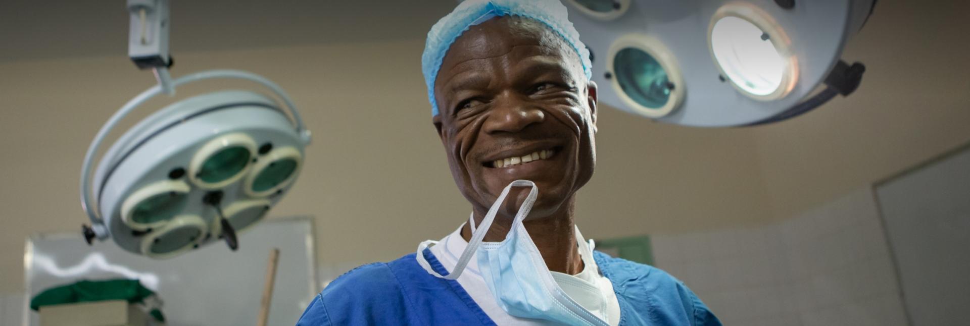 A man surrounded by medical equipment and dressed in a surgical gown smiles and holds a mask by his teeth.