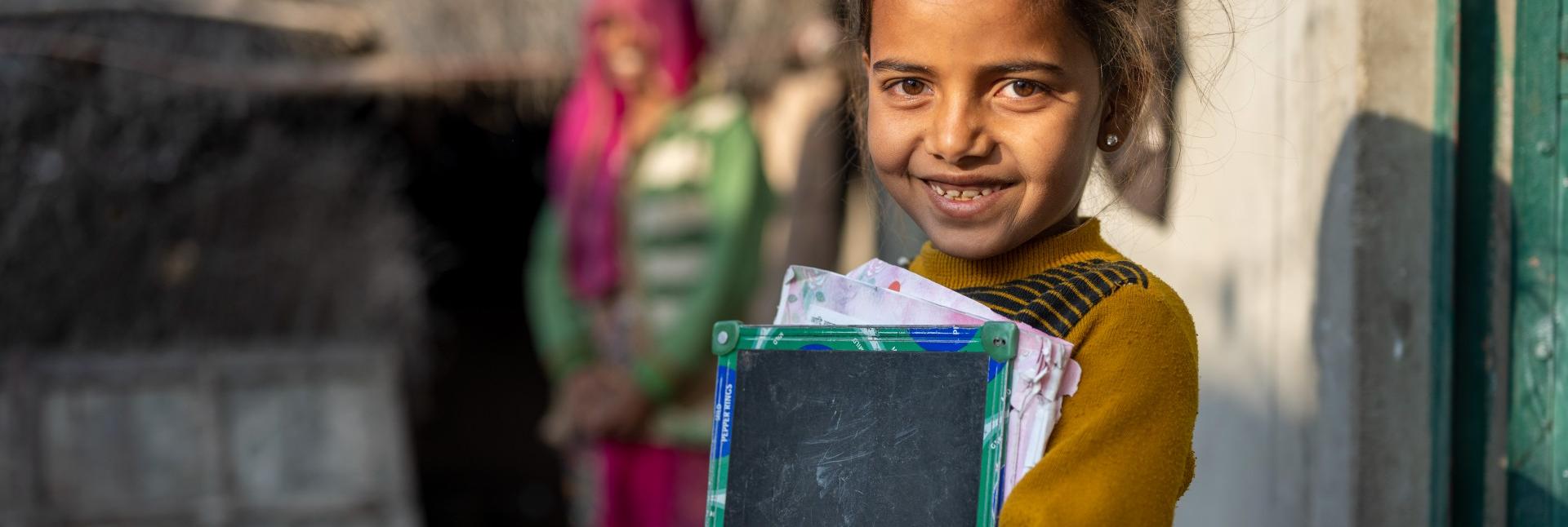 A young girl holds her school-work in her hands, in front of a house, at dusk.